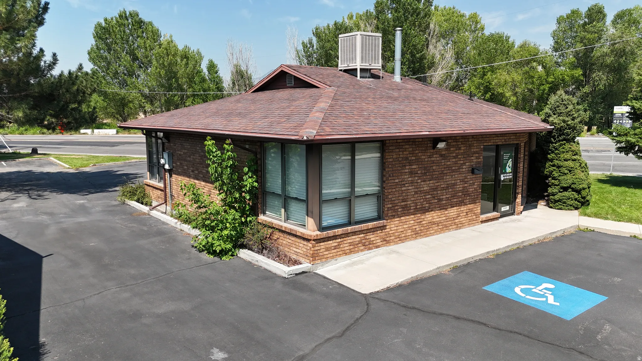 Brick exterior with a peaked shingle roof, multiple windows, and an asphalt parking area