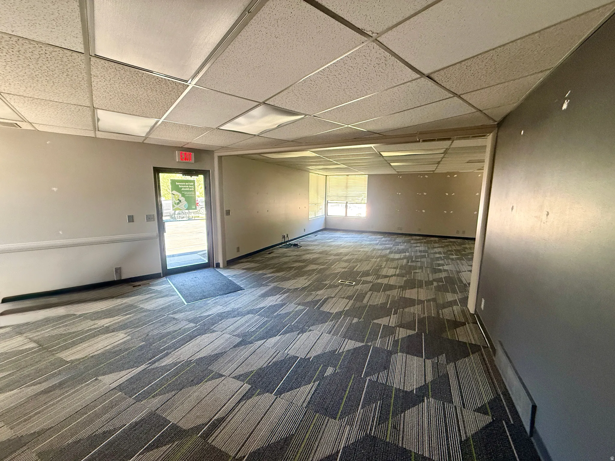 Spacious open-concept interior featuring modular carpet tile flooring, drop ceiling with integrated lighting, multiple windows with blinds, and a glass-paneled entry door