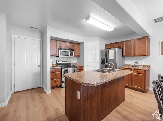 Kitchen featuring wood-finish flooring, stainless steel appliances, a center island with a built-in sink, wood cabinetry, and a tile backsplash