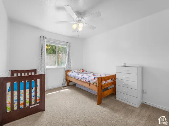 Bright bedroom featuring carpeted flooring, ceiling fan with integrated lighting, large window with natural light, wood-finish bed frame, and white drawer dresser for storage