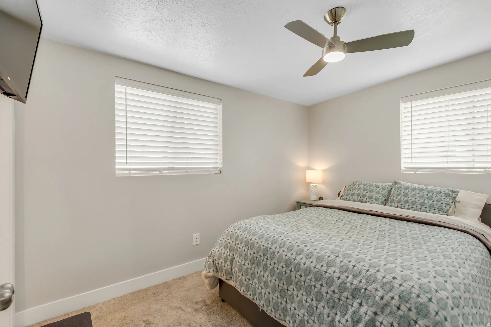 Neutral wall paint, ceiling fan with integrated lighting, two windows with horizontal blinds, and carpeted flooring