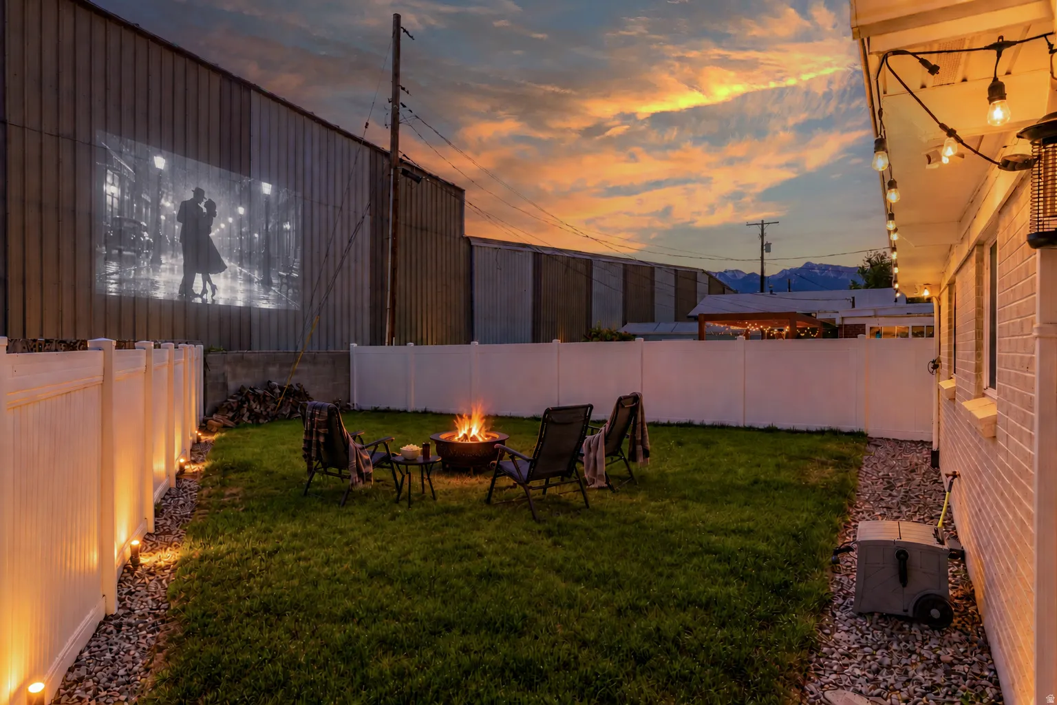 Fenced backyard featuring a manicured lawn, fire pit, string lighting, and exterior projection space