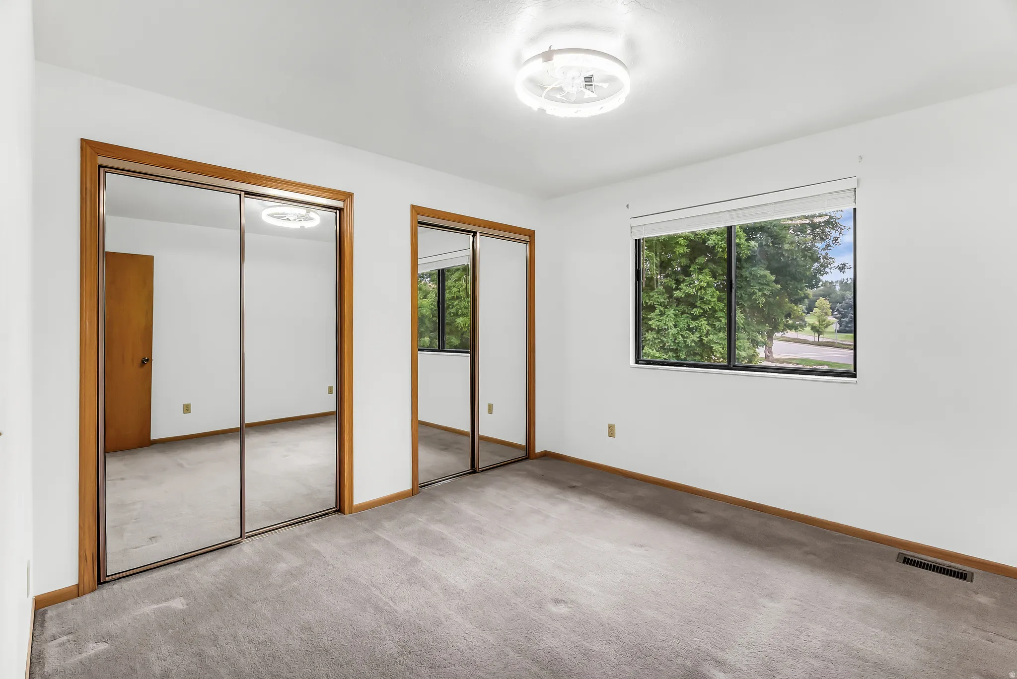 Light-colored carpeting, two mirrored closet doors with wood trim, a single window, and a ceiling-mounted light fixture