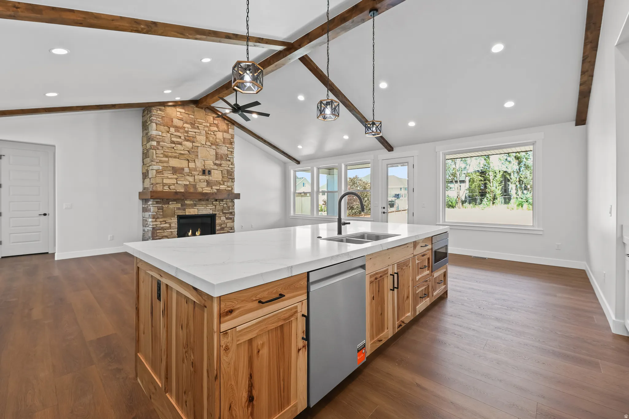Vaulted ceilings with exposed wood beams, stone fireplace, kitchen island with built-in sink and dishwasher, wood-finish flooring, and multiple windows