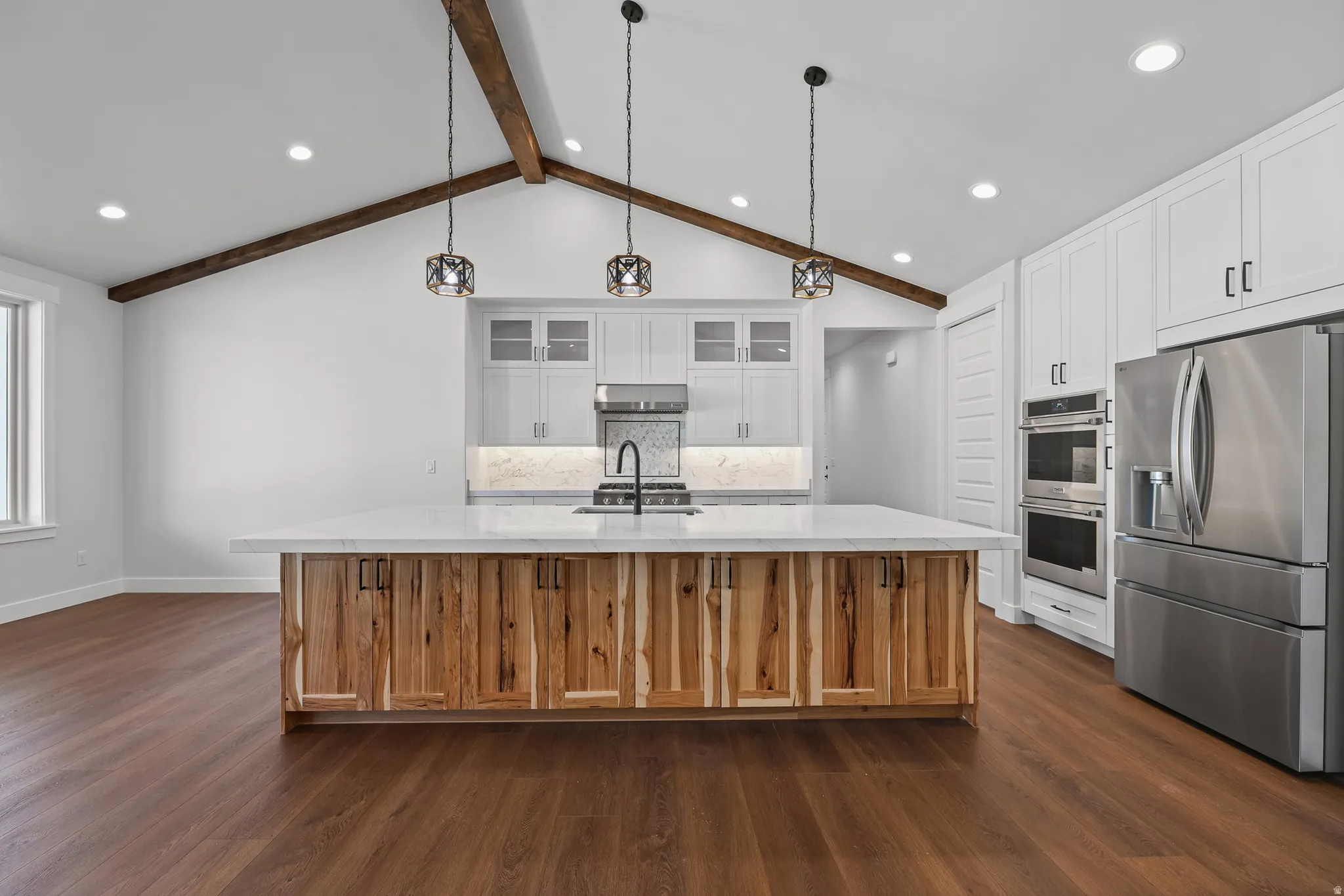 Vaulted ceiling with exposed wood beams, wood-finish flooring, stainless steel appliances, large kitchen island with wood-paneled cabinetry, and recessed lighting