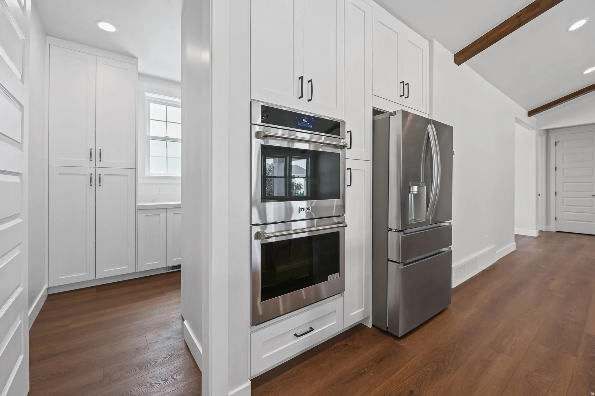 Stainless steel double ovens, stainless steel refrigerator, white shaker cabinetry, wood-finish flooring, and vaulted ceiling with exposed beams