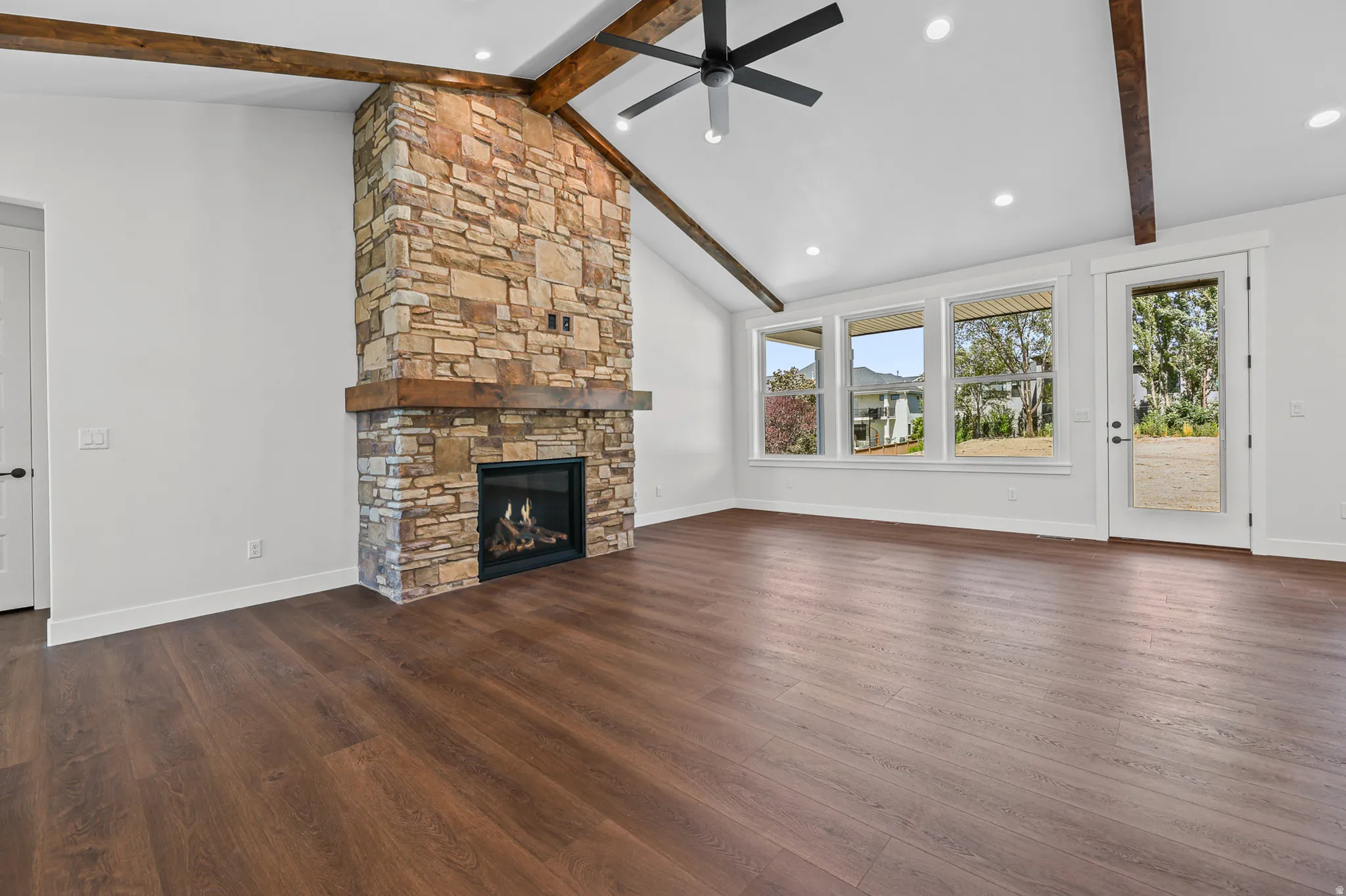 Vaulted ceiling with exposed wood beams, stone-clad fireplace with wood mantel, wood-finish flooring, recessed lighting, and multiple windows