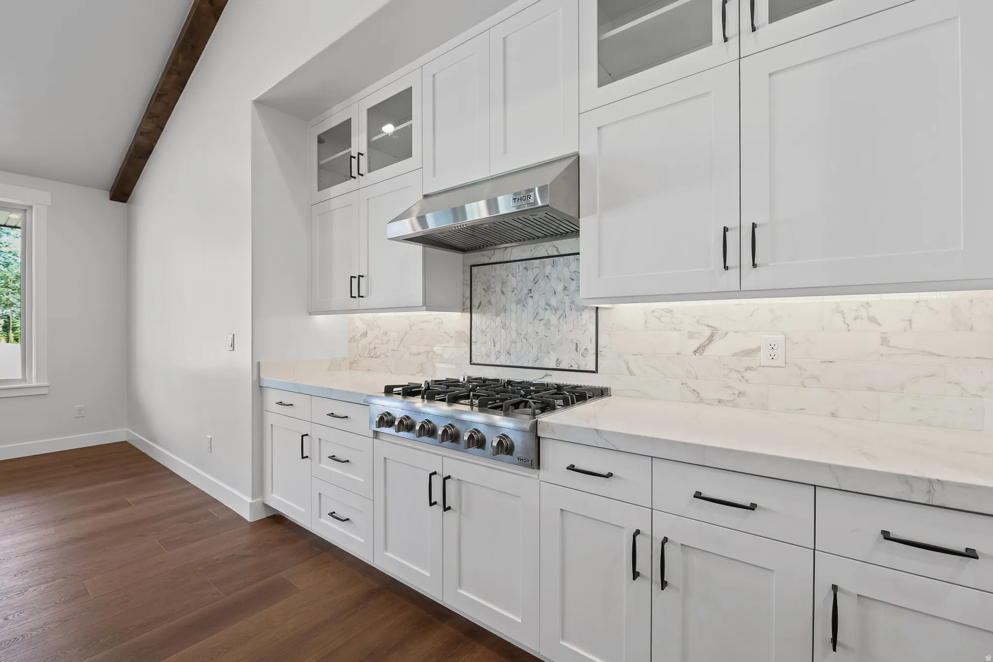 White flat-panel cabinetry, stainless steel range with overhead hood, integrated under-cabinet lighting, wood-finish flooring, and a visible wood beam