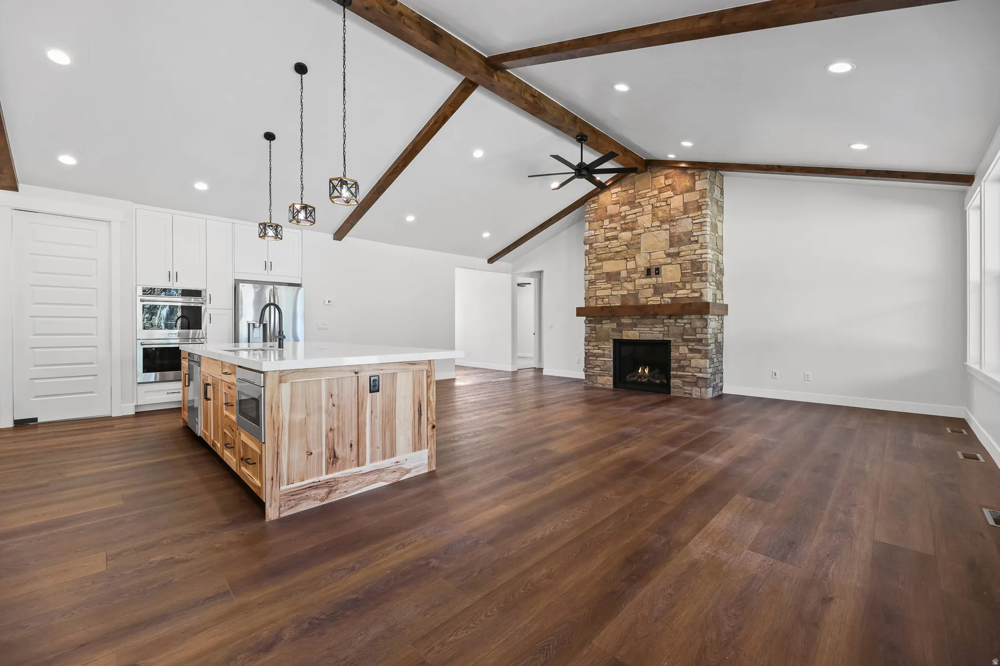 Vaulted ceiling with exposed wood beams, wood-finish flooring, stone-clad fireplace with wood mantel, kitchen island with built-in appliances, and white cabinetry