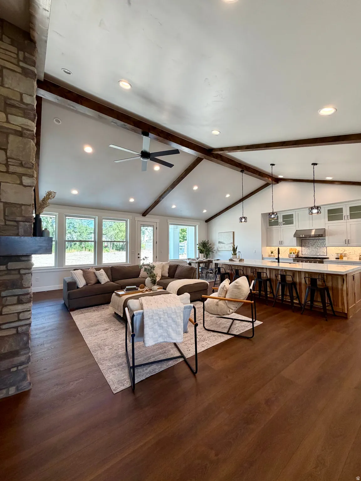 Vaulted ceiling with exposed wood beams, wood-finish flooring, stone-clad fireplace, recessed lighting, and a kitchen island with seating