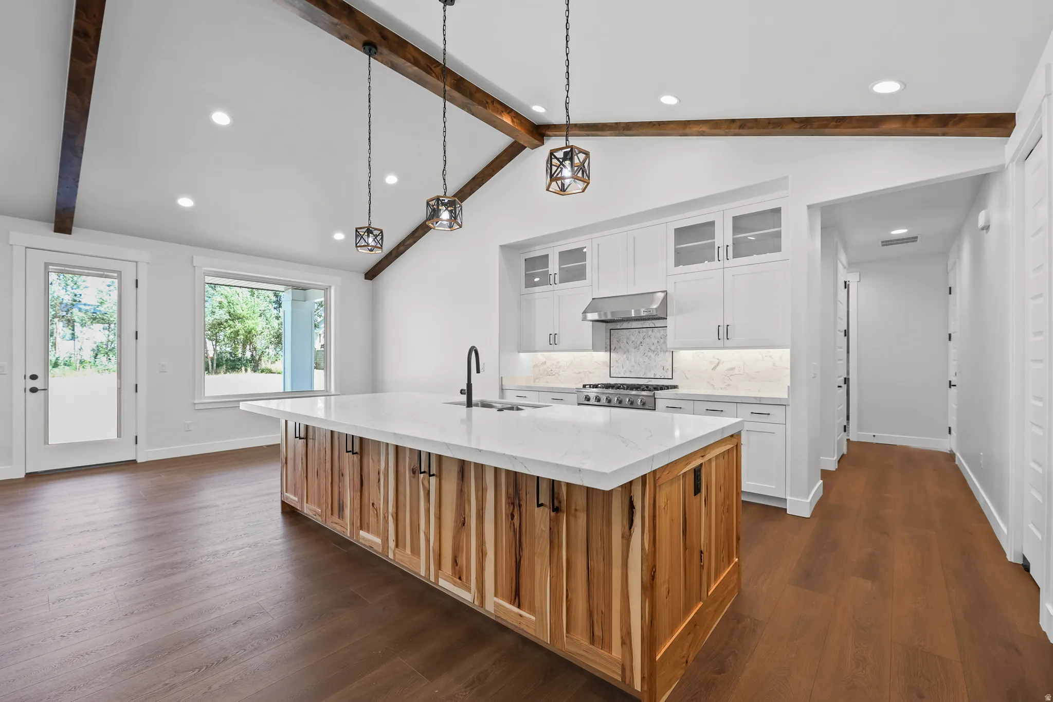 Vaulted ceiling with exposed wood beams, wood-finish flooring, white cabinetry, a large kitchen island with a light-toned countertop, and recessed lighting