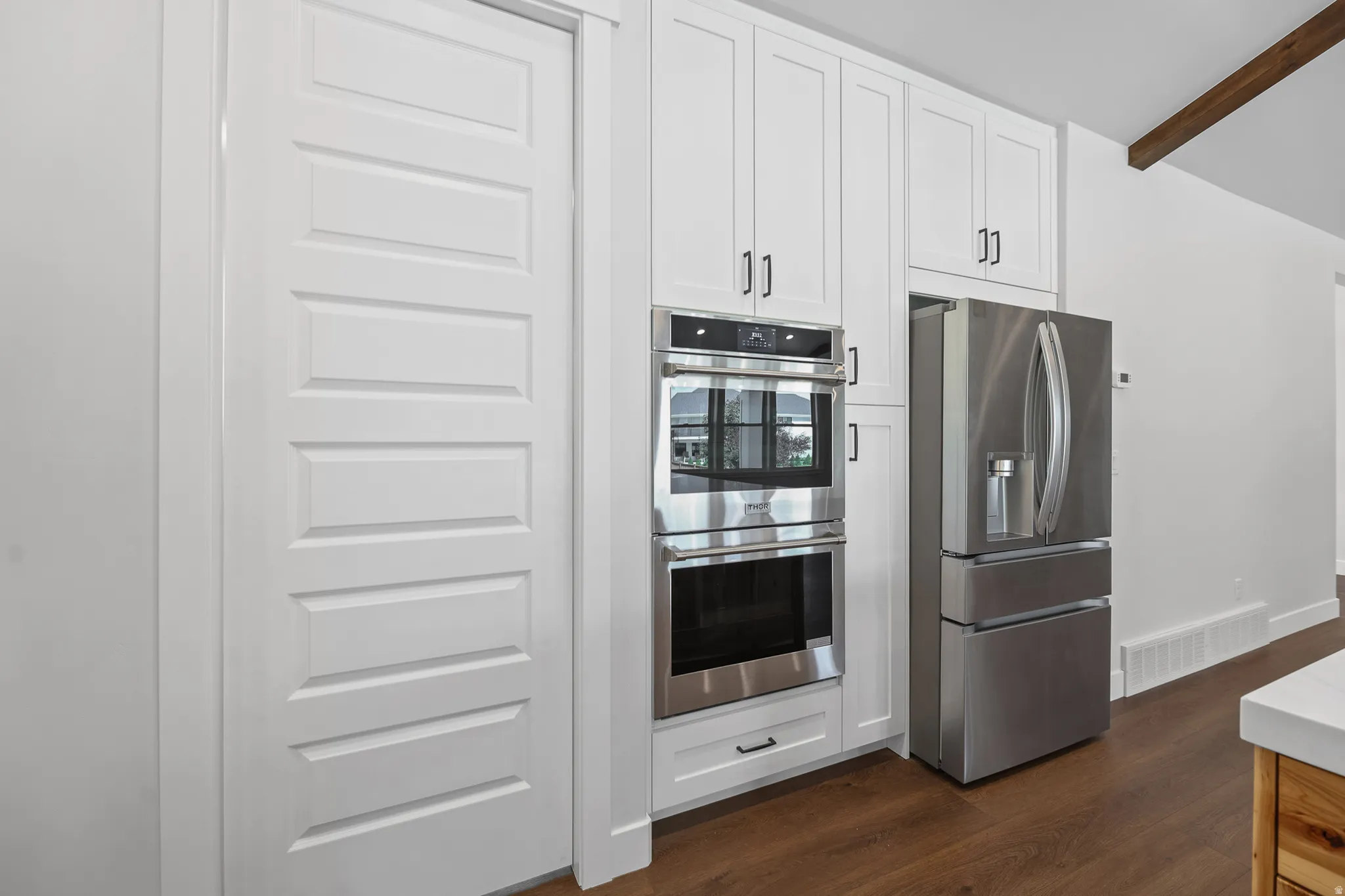 White shaker cabinetry with black hardware, stainless steel double wall ovens, stainless steel refrigerator, and wood-finish flooring