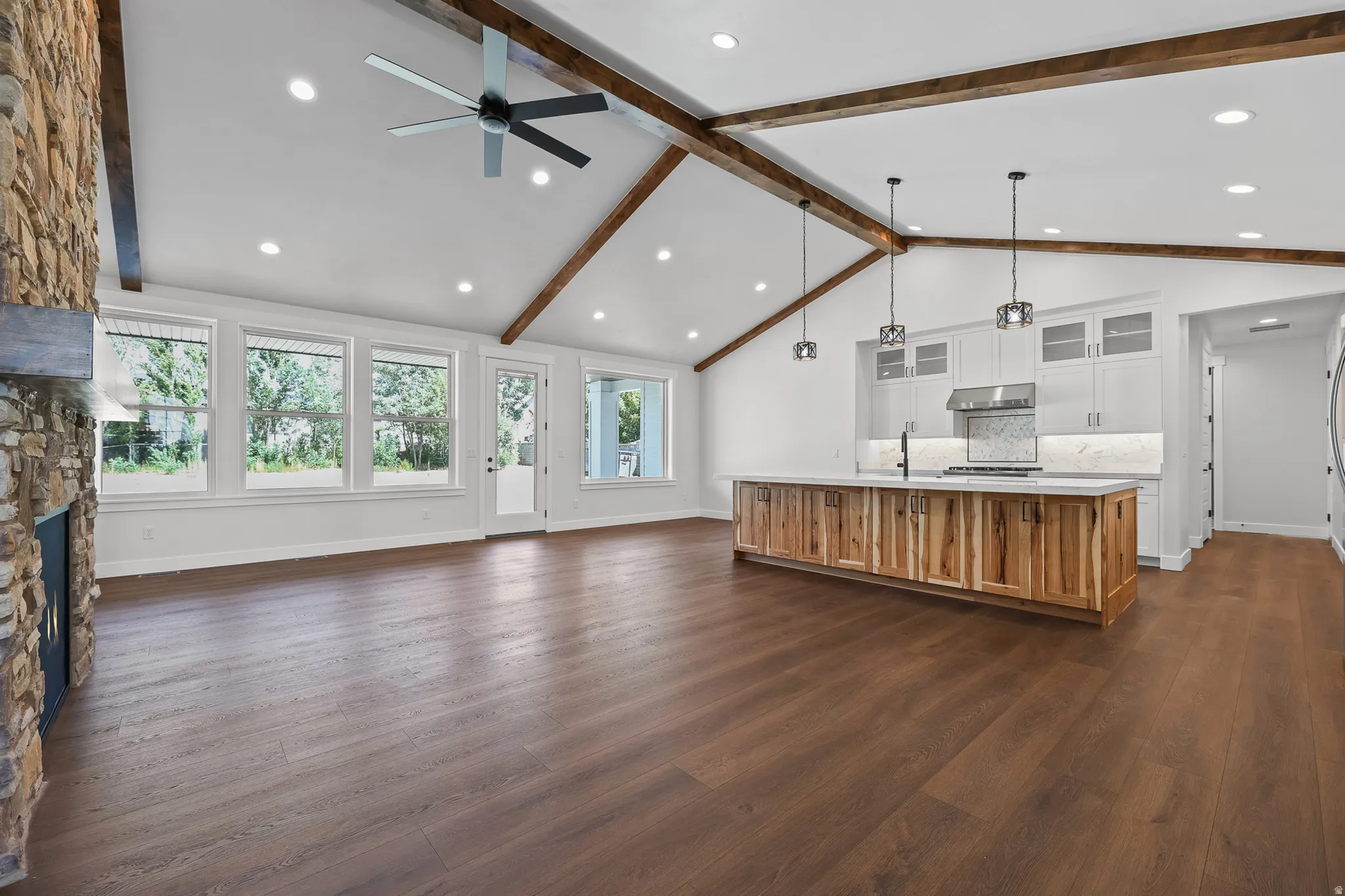 Vaulted ceiling with exposed wood beams, wood-finish flooring, kitchen island with wood-finish cabinetry, stone fireplace, and recessed lighting