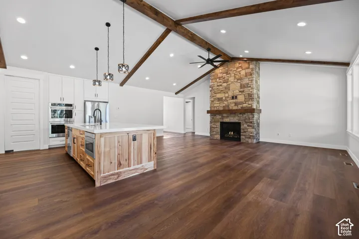 Vaulted ceiling with exposed wood beams, wood-finish flooring, stone-clad fireplace with wood mantel, kitchen island with built-in appliances, and white cabinetry