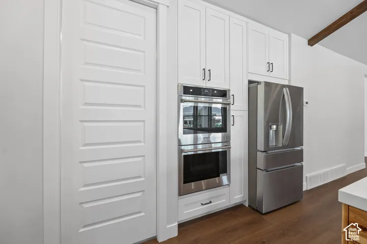 White shaker cabinetry with black hardware, stainless steel double wall ovens, stainless steel refrigerator, and wood-finish flooring