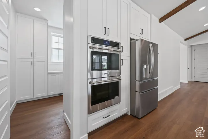 Stainless steel double ovens, stainless steel refrigerator, white shaker cabinetry, wood-finish flooring, and vaulted ceiling with exposed beams
