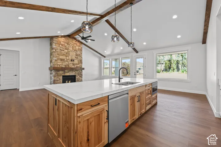 Vaulted ceilings with exposed wood beams, stone fireplace, kitchen island with built-in sink and dishwasher, wood-finish flooring, and multiple windows