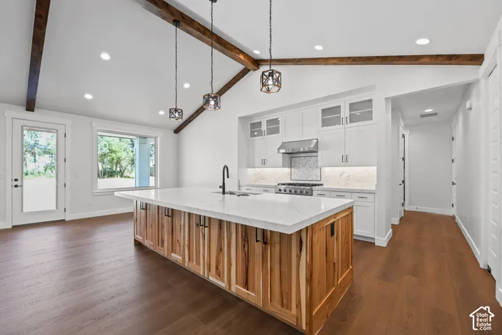 Vaulted ceiling with exposed wood beams, wood-finish flooring, white cabinetry, a large kitchen island with a light-toned countertop, and recessed lighting
