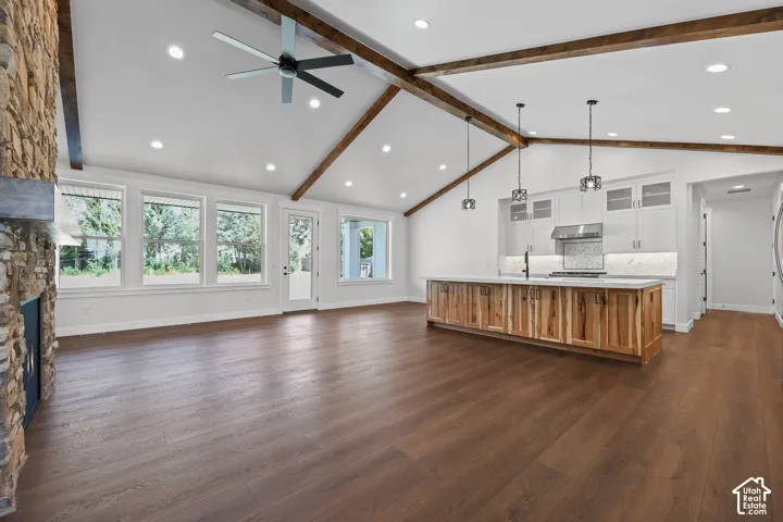 Vaulted ceiling with exposed wood beams, wood-finish flooring, kitchen island with wood-finish cabinetry, stone fireplace, and recessed lighting