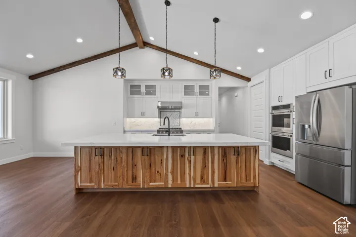 Vaulted ceiling with exposed wood beams, wood-finish flooring, stainless steel appliances, large kitchen island with wood-paneled cabinetry, and recessed lighting