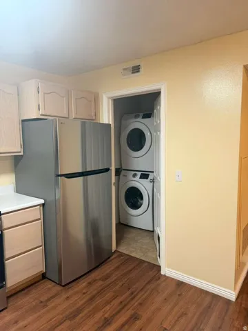 Stainless steel refrigerator, light-toned cabinetry, white countertops, wood-finish flooring, and a stacked washer and dryer in a dedicated alcove