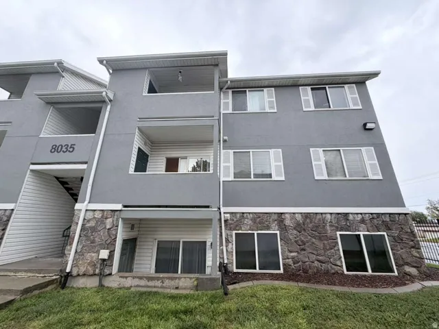 Multi-level building with gray stucco siding, stone veneer base, white window shutters, and white rain gutters