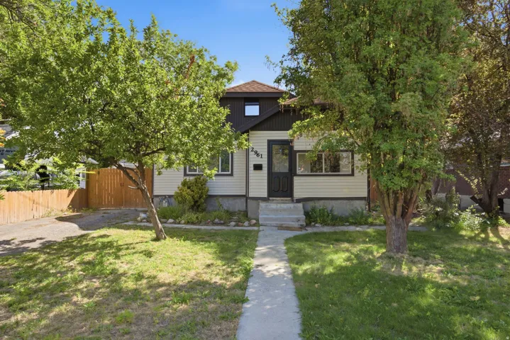 Light-colored siding exterior with dark trim accents, concrete walkway, mature trees, and a wooden fence