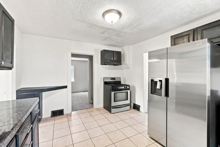 Stainless steel refrigerator, tile flooring, built-in cabinetry with dark finishes, gas range, and wall-mounted light fixture