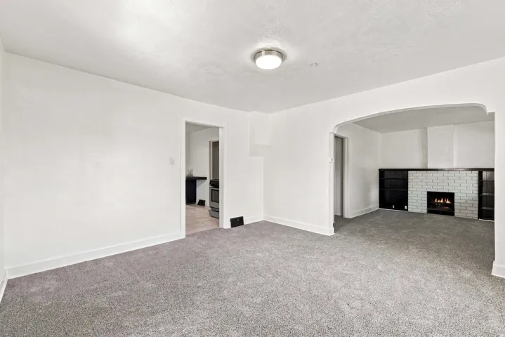 Plush gray carpeting throughout, arched doorway entry, and a white brick fireplace with built-in dark cabinetry