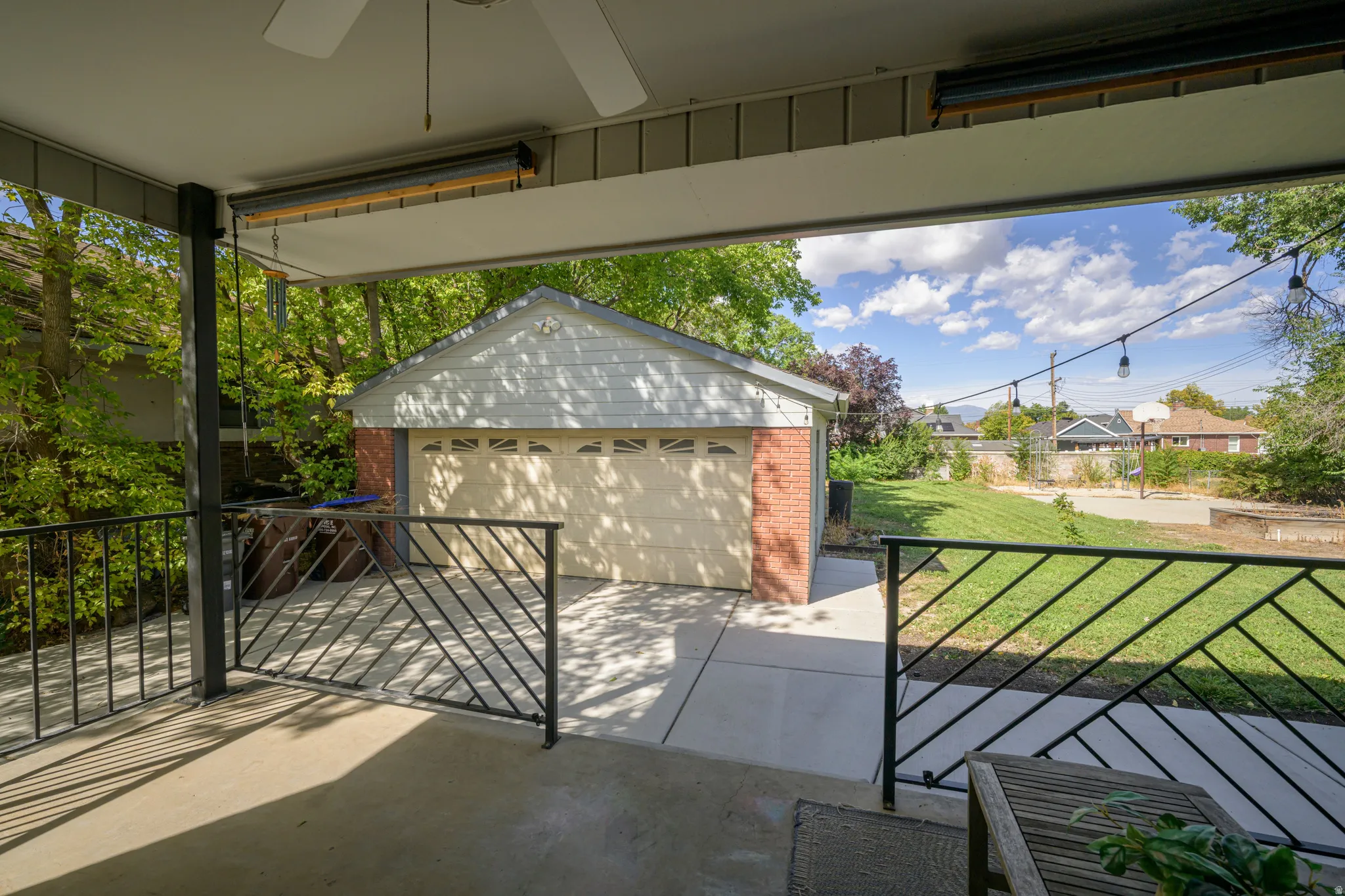 Covered patio with ceiling fan, black metal railing, and string lights, overlooking a detached garage with brick siding and a concrete driveway