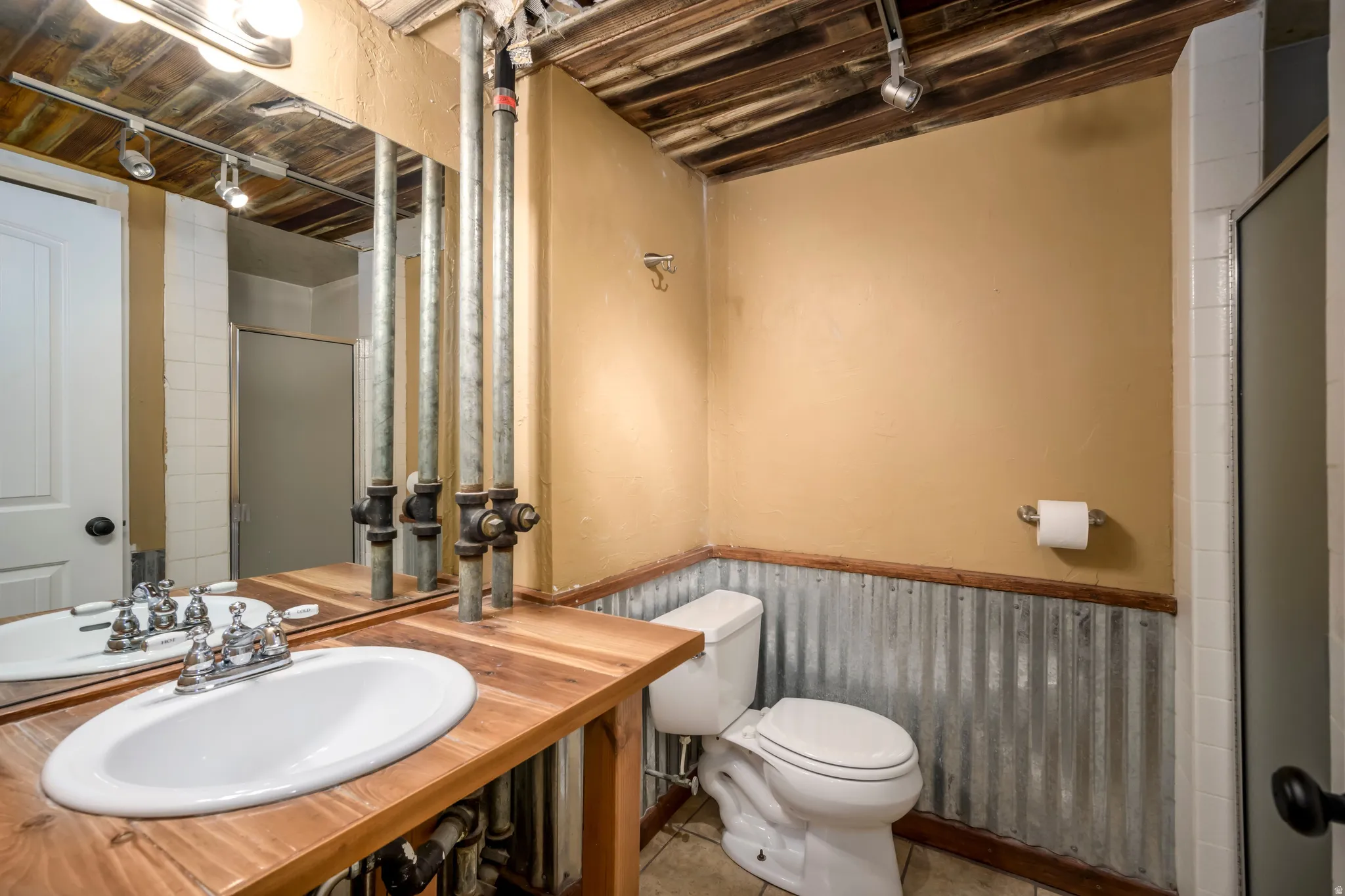 Wood-finish vanity with an oval drop-in sink, corrugated metal wainscoting, exposed wood beam ceiling, and a shower enclosure