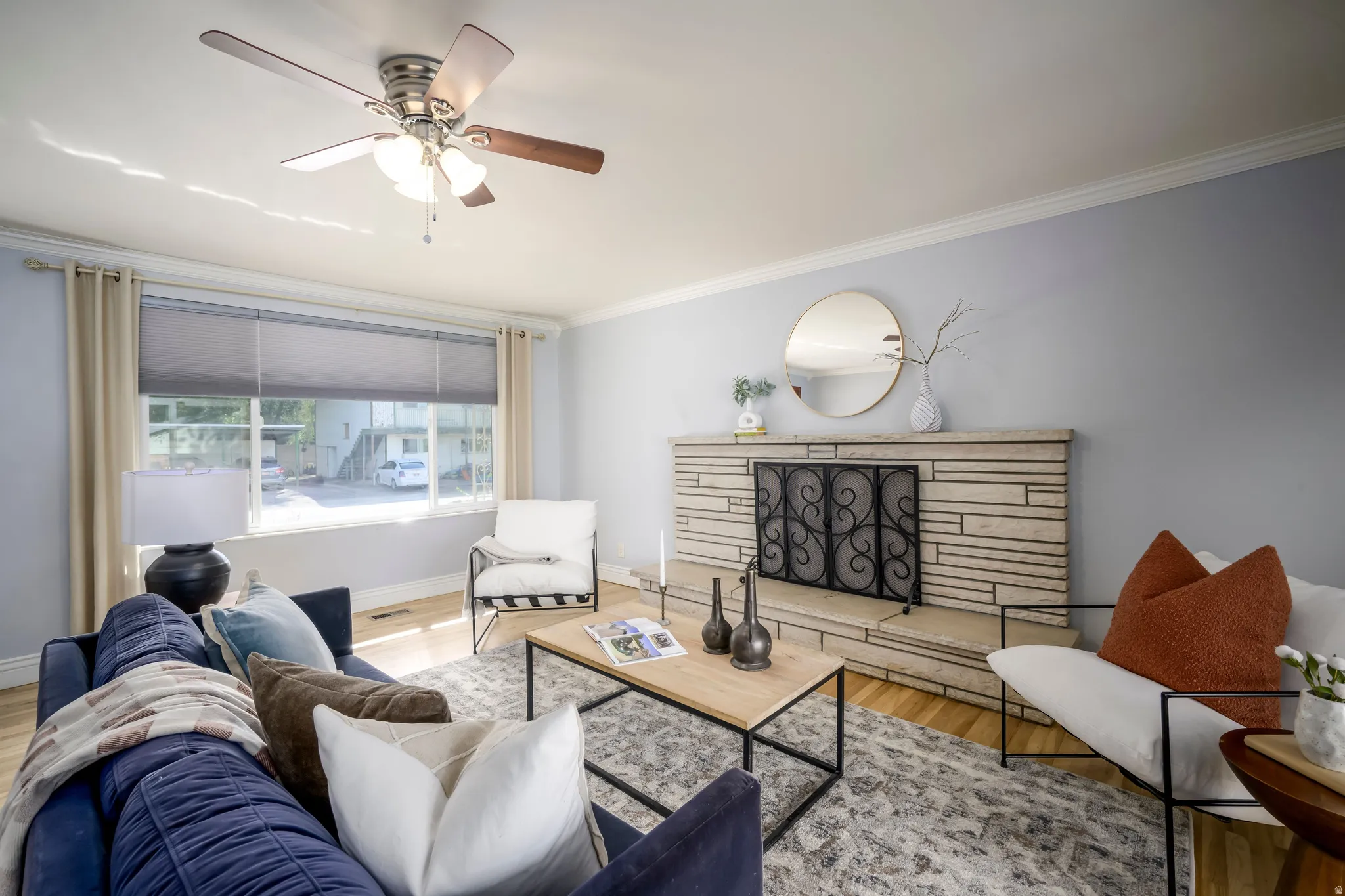 Living area featuring wood-finish flooring, a ceiling fan with integrated lighting, a stone-clad fireplace with an ornate screen, and crown molding