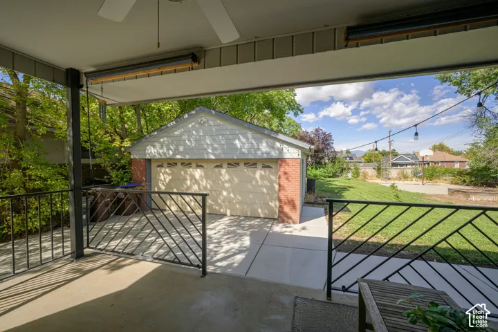 Covered patio with ceiling fan, black metal railing, and string lights, overlooking a detached garage with brick siding and a concrete driveway