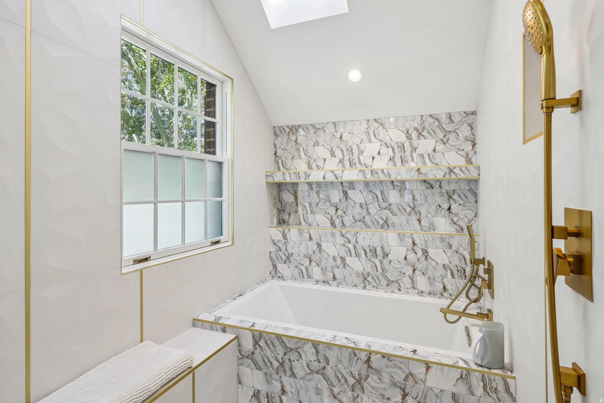 Built-in bathtub with mixed-pattern tile surround, recessed wall shelving, gold-finish fixtures, and a skylight