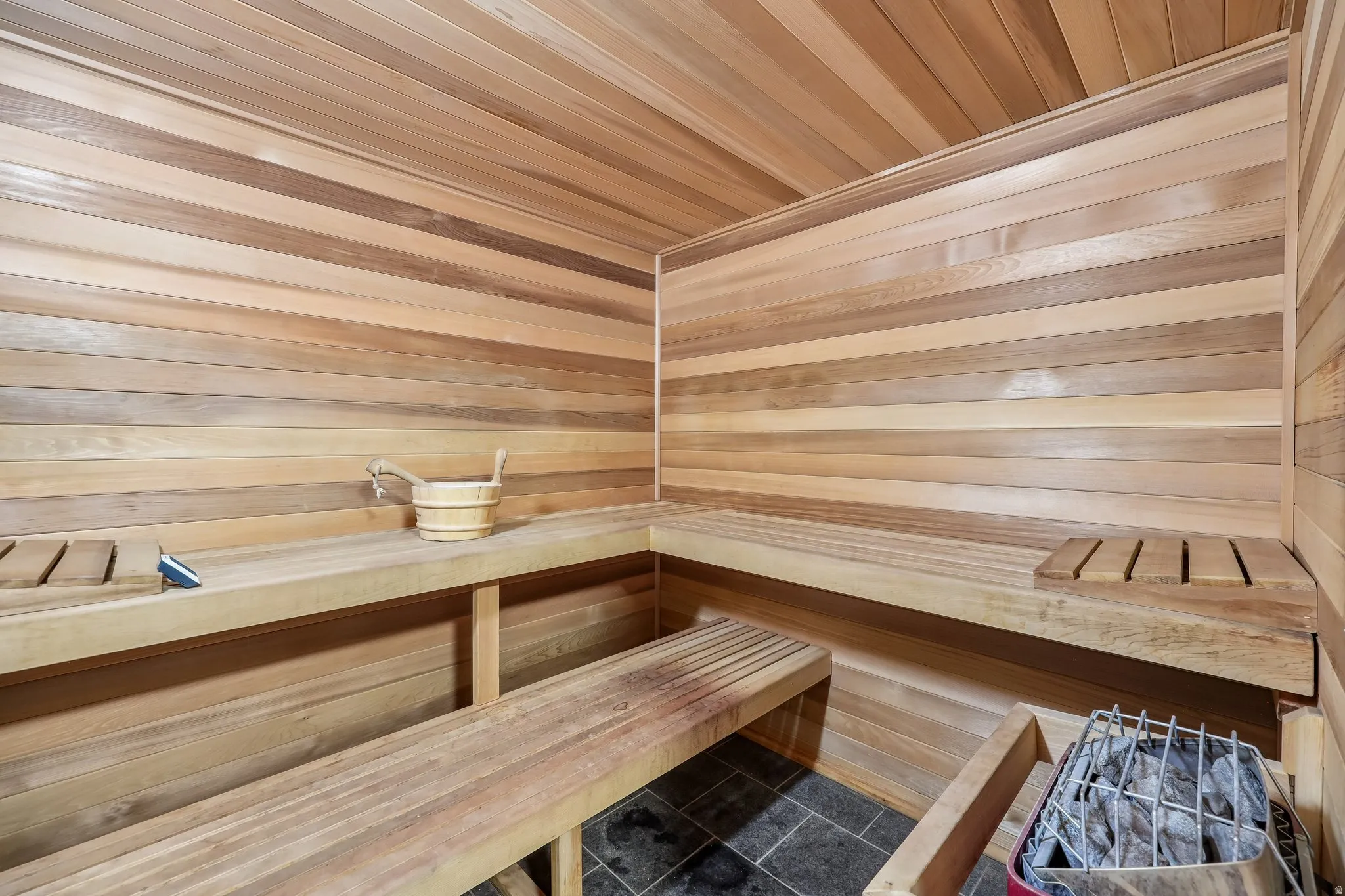 Wood-paneled sauna with built-in tiered benches, dark tile flooring, and a stone heater