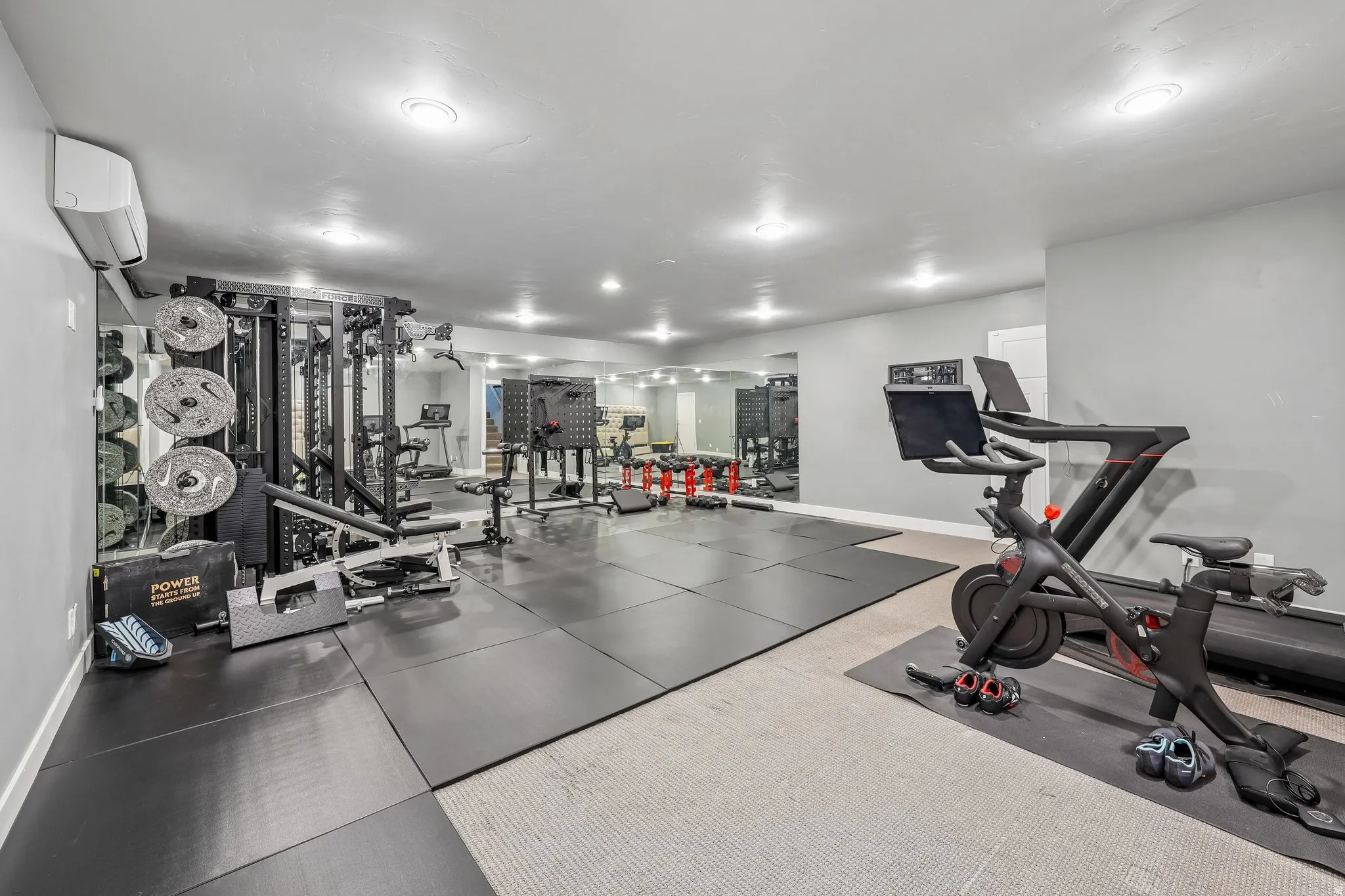 State-of-the-art home fitness center featuring professional-grade multi-station strength training equipment, stationary cycling bike with digital console, dark tile flooring throughout, recessed LED ceiling lighting, and ample open space for functional tr