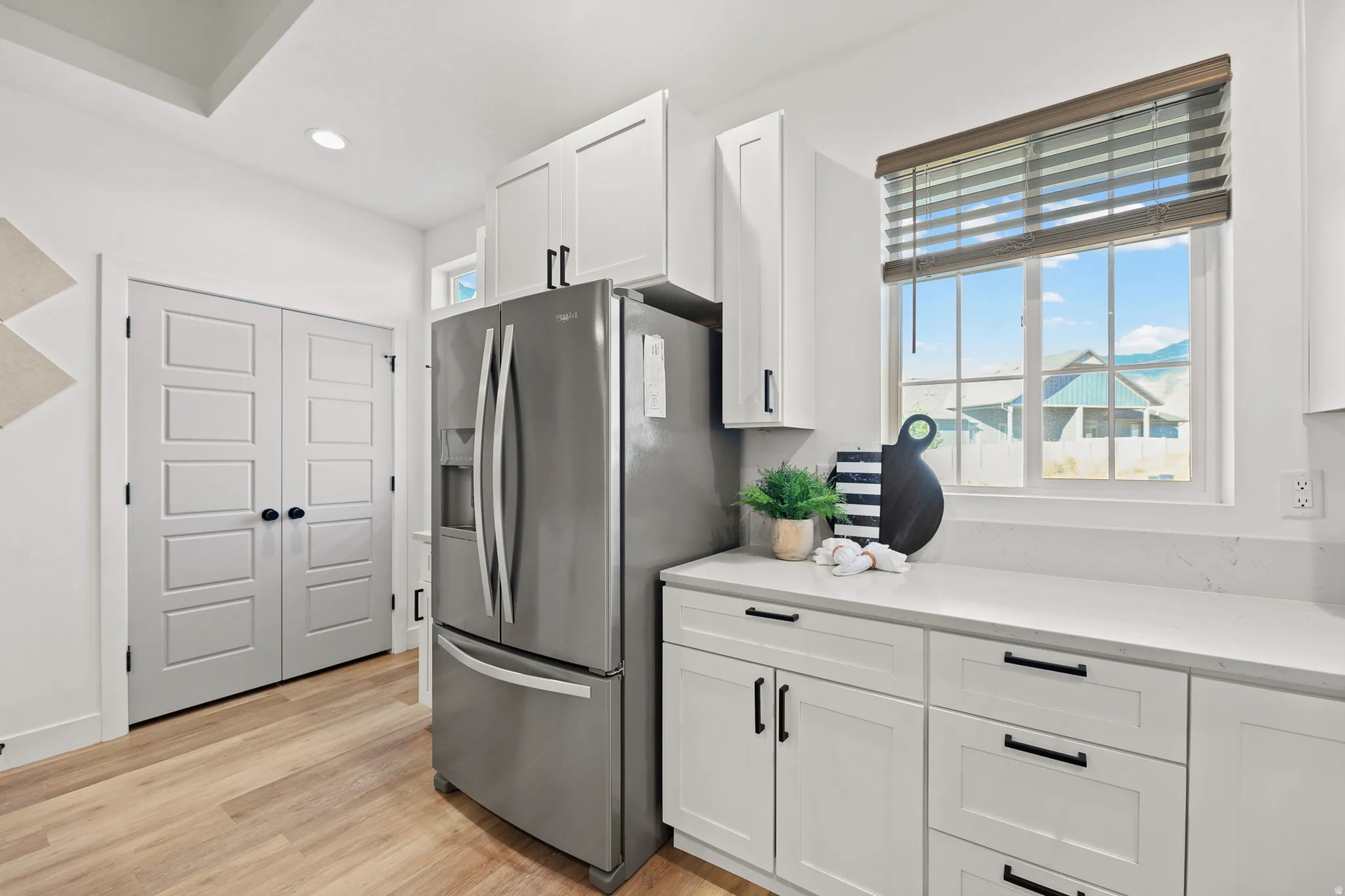 Stainless steel refrigerator, white shaker-style cabinetry, white countertops, wood-finish flooring, and a large window with blinds