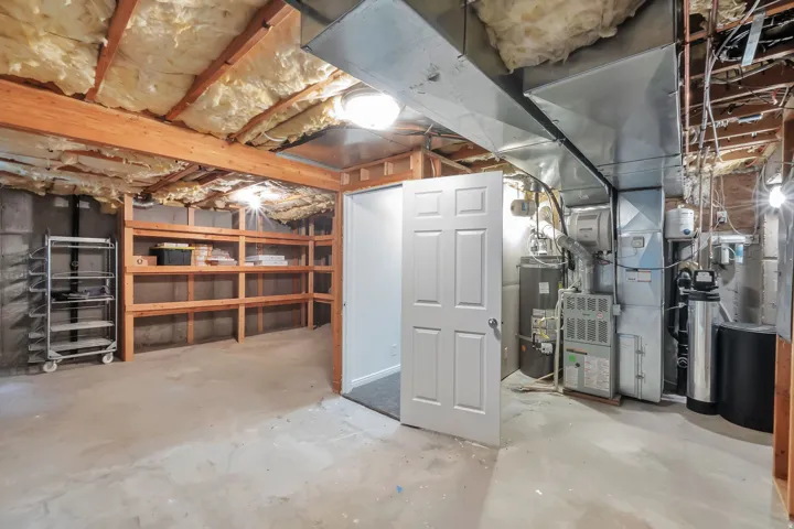 Unfinished basement featuring exposed wood beams, concrete flooring, built-in wooden shelving, and utility systems