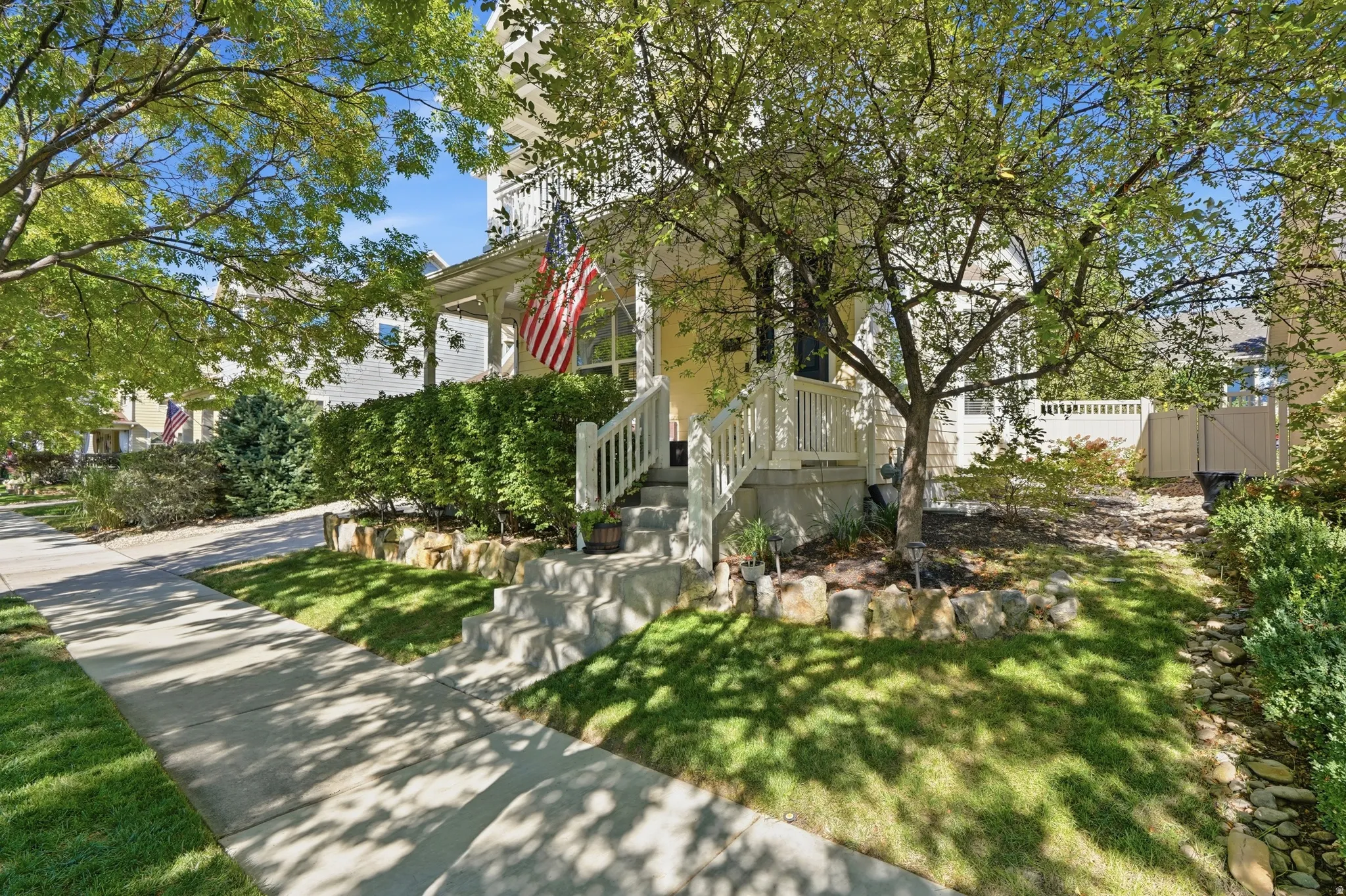 Front porch with white railings, concrete steps, mature trees, manicured lawn, and established hedges