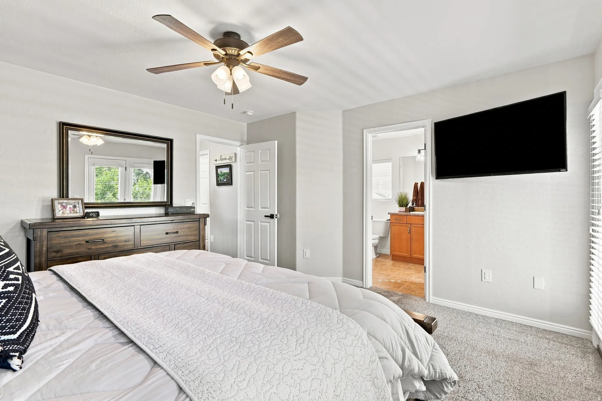 Carpeted flooring, ceiling fan with integrated lighting, wall-mounted television, and an adjacent bathroom with a wood-finish vanity