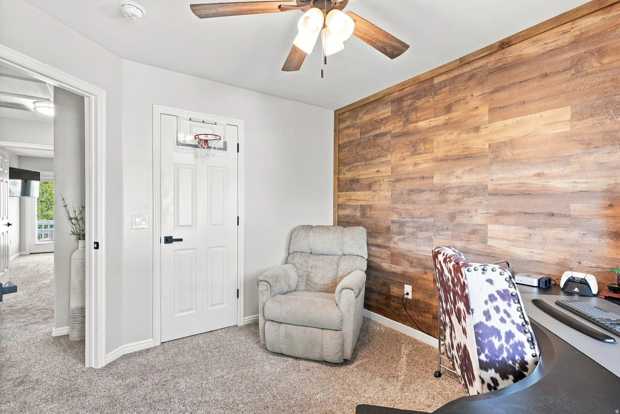 Accent wall with wood-finish paneling, ceiling fan with light fixture, light gray carpeting, and paneled interior door with matte black hardware