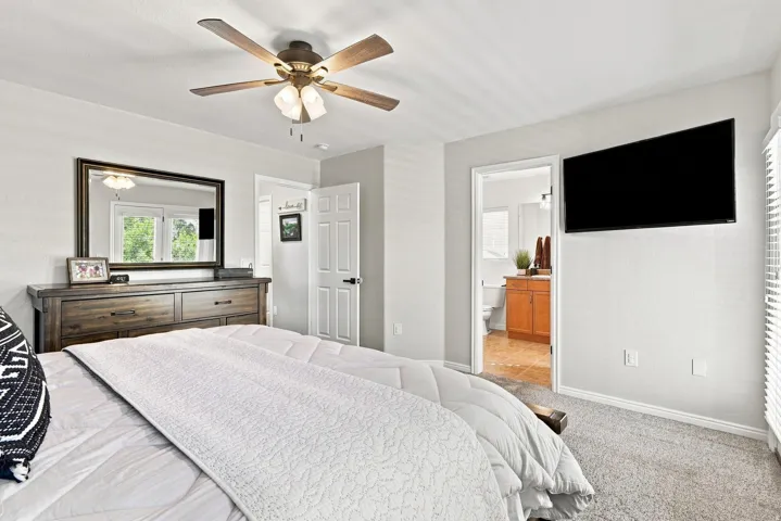 Carpeted flooring, ceiling fan with integrated lighting, wall-mounted television, and an adjacent bathroom with a wood-finish vanity