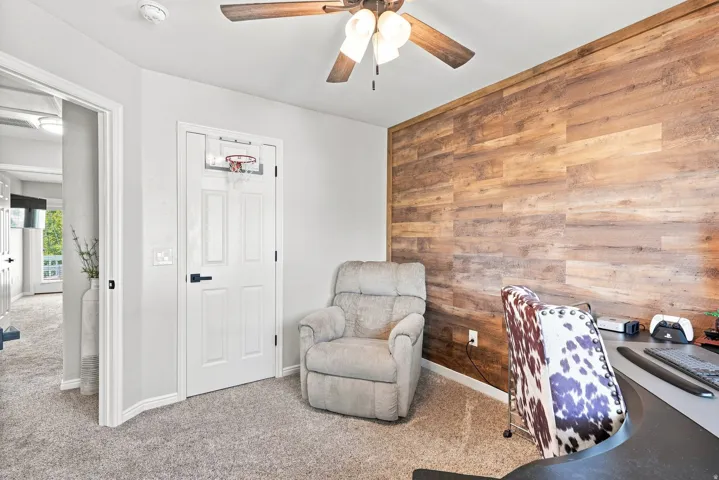 Accent wall with wood-finish paneling, ceiling fan with light fixture, light gray carpeting, and paneled interior door with matte black hardware