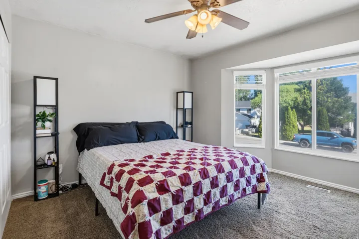 Bay window with multiple panes, ceiling fan light fixture, light gray wall paint, and textured carpet flooring