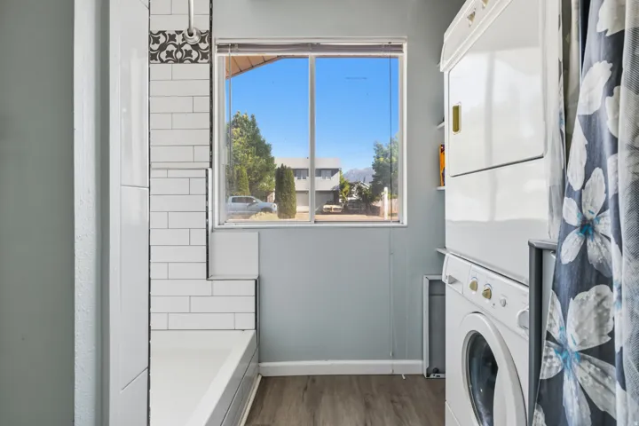 White stacked washer and dryer unit, wood-finish flooring, white subway tile surround, decorative tile accent, and a single window