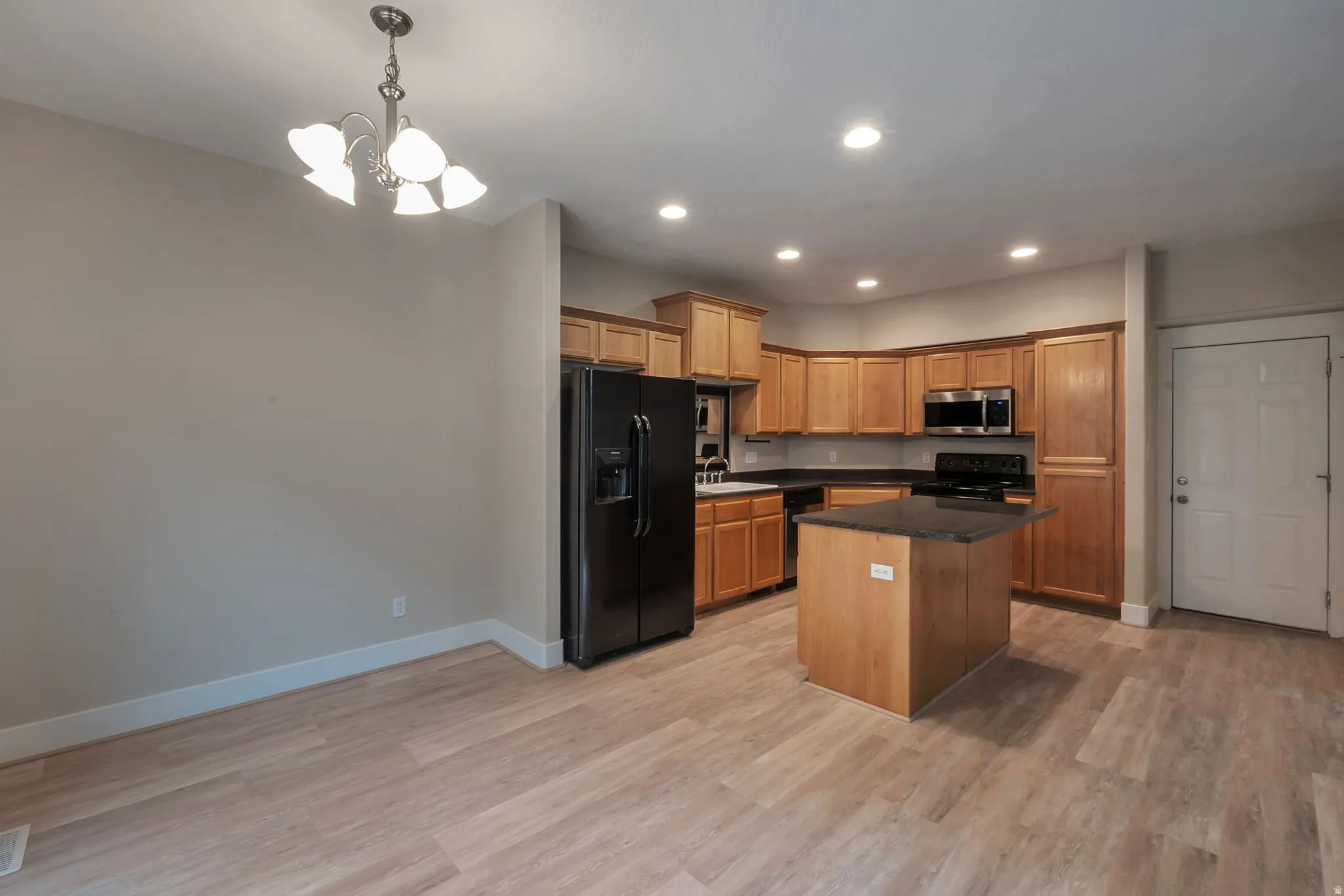 LVP hardwood-style flooring throughout, kitchen with wood cabinetry and black appliances, kitchen island with dark countertop, recessed lighting, and chandelier
