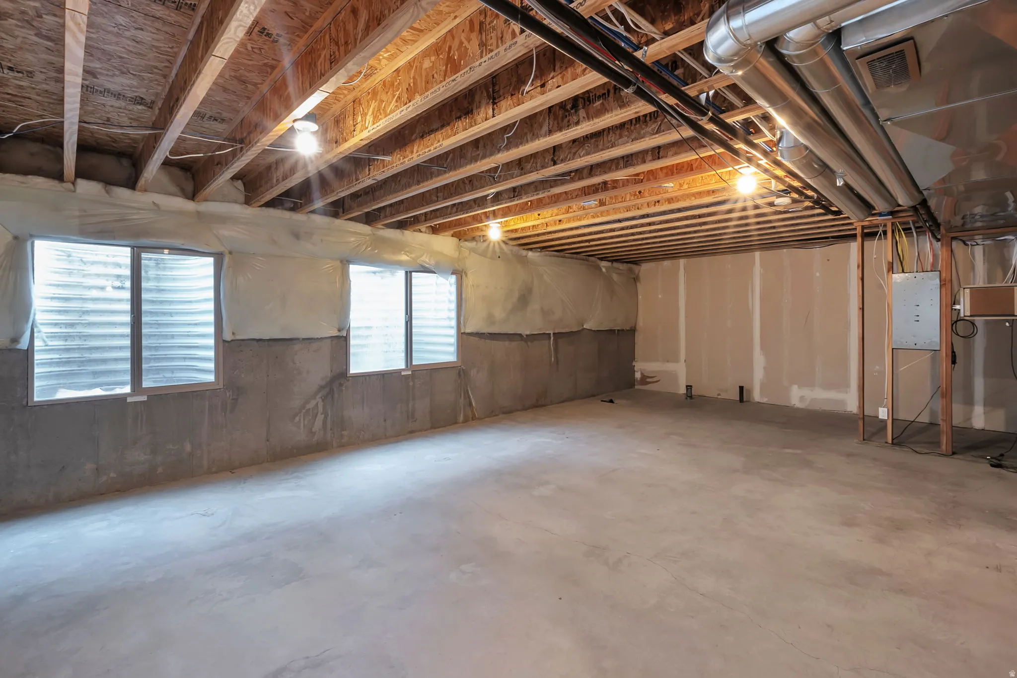 Unfinished basement featuring exposed wood joists, concrete flooring, concrete walls, three basement windows, and visible utility conduits