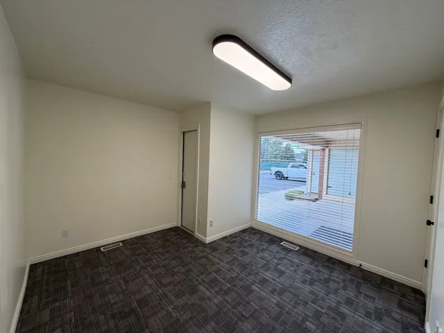 Room featuring a large window with blinds, dark patterned carpeting, and an overhead linear light fixture