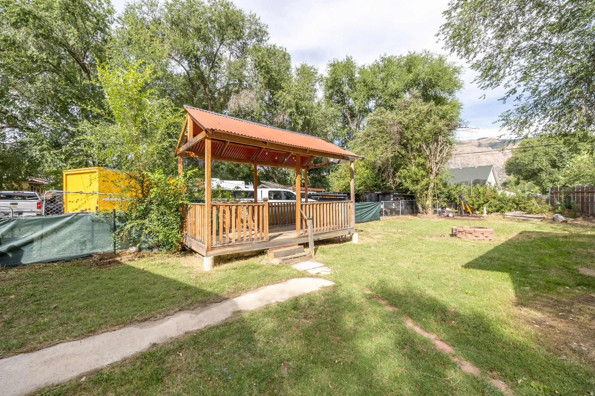 Expansive backyard featuring a covered wooden gazebo with a corrugated metal roof, a concrete walkway, and a brick fire pit