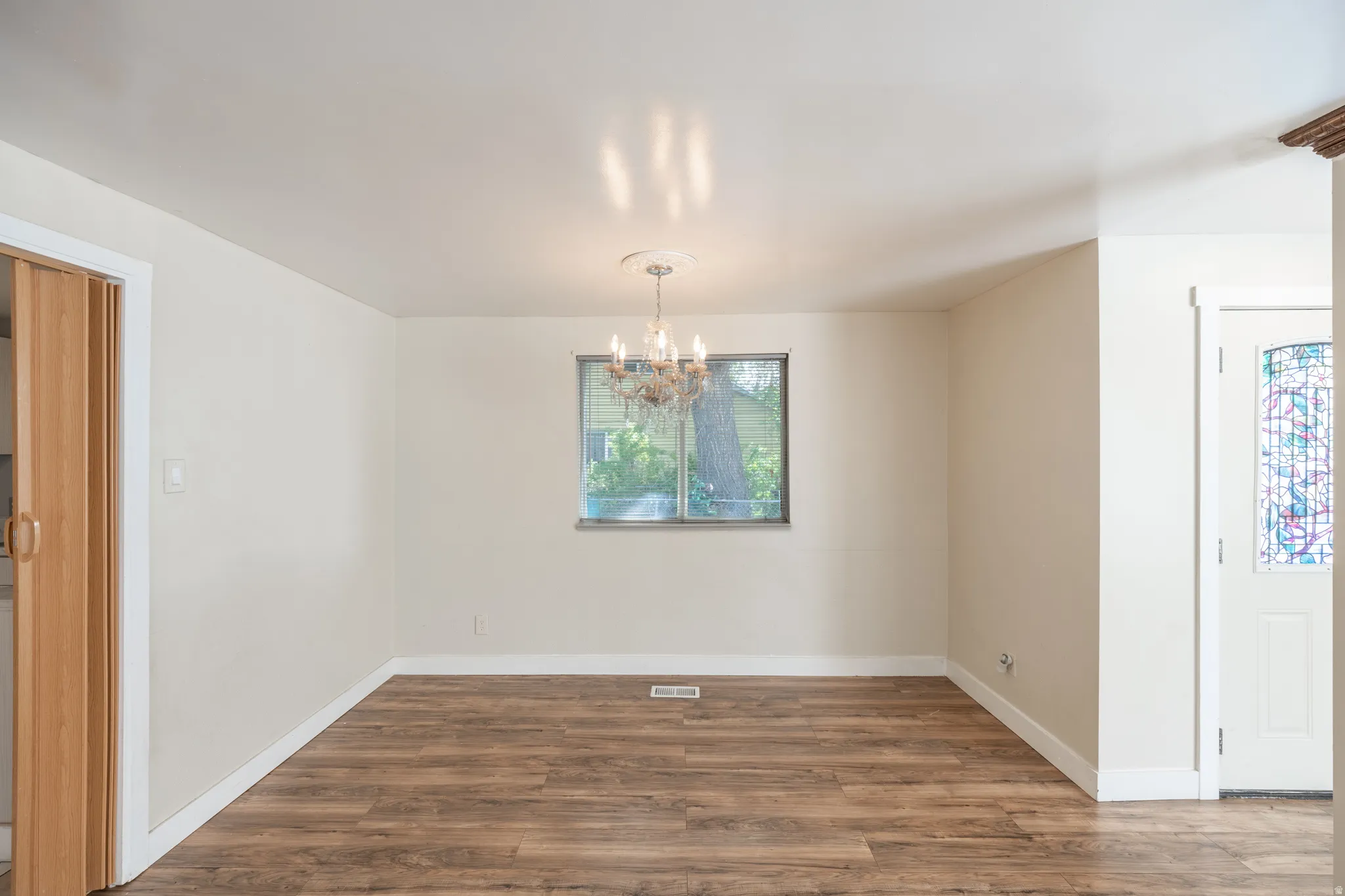 Wood-finish flooring, central window, ceiling-mounted chandelier with medallion, white trim, and a decorative stained glass door